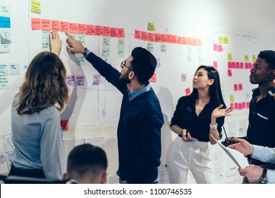 Team of multicultural young people pointing on wall with glued colorful paper notes with foreign words during productive lesson.Diverse group of male and female employees in formal wear using stickers