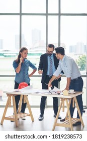 Team Of Multiethnic Architects Working On Construction Plans In Meeting Room. Engineers Discussing On Project In Office. Mature Businessman And Woman Standing Around Table Working On Blueprint.