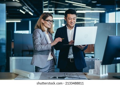 A Team Of Multiracial Business Architects And Designers Discuss Creating A Project Draft Drawing By Looking At Laptop Monitor Screen A Computer Desk. Asian Male And Female Developers Together Teamwork