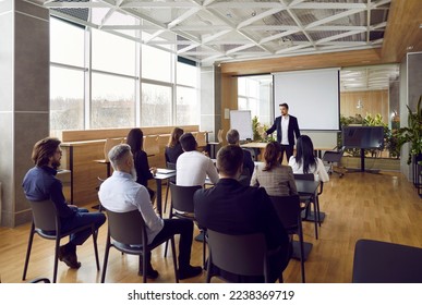 Team of people having class with business trainer. Group of male and female employees sitting at desks in modern office and listening to lecture by experienced teacher sharing knowledge and guidance
