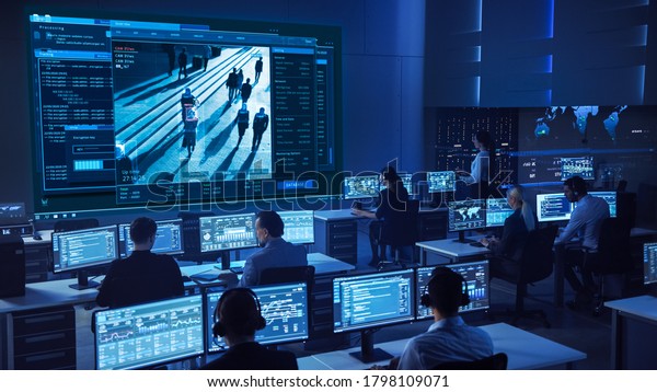 Team of Professional Cyber Security Data
Science Engineers Work on Surveillance Tracking Shot of People
Walking on City Streets. Big Dark Control and Monitoring Room with
Computer Displays.