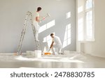 Team of professional painters in white clothes painting a grey wall in empty room with paint roller. Building contractors doing repair renovation in client house. Repair and renovating concept.