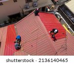 A team of roofers or carpenters installs new corrugated metal roofing with ribbed sheet design on a two storey house. Using a safety harness for protection. Replacing older roof.