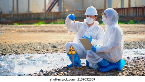 Team Scientist In Protective Suit, Mask And Gloves Collect Sample Of Fish And Dirty Water From Factory  Took A Sample Of Waste Water For Analysis. Pollution And Environment Problems