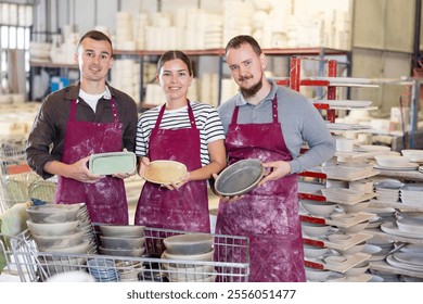 Team of smiling professional, skilled artisans, young woman and two men in stained burgundy aprons, presenting handcrafted ceramic dishes in collaborative pottery workshop - Powered by Shutterstock