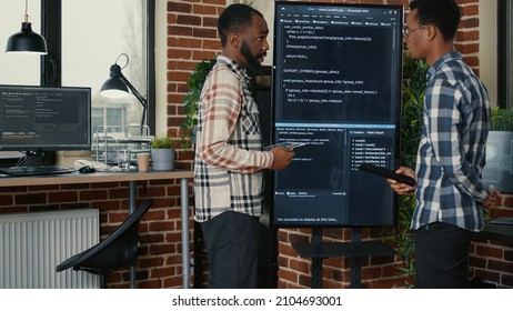 Team of software engineers leaving after analyzing source code on wall screen tv comparing errors using digital tablet. Coders passing coworker programmer writing code for artificial intelligence.