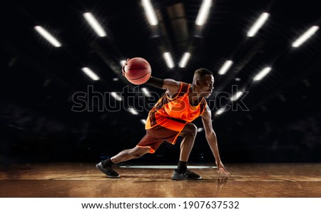 Team supporting. African-american young basketball player in action and motion in flashlights over dark gym background. Concept of sport, movement, energy and dynamic, healthy lifestyle. Arena's