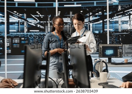 Team of technicians using tablet in server farm, analyzing data. Women working in data center examining infrastructure using device, ensuring system integrity and security