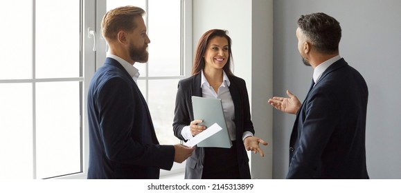 Team Of Three Cheerful Friends And Coworkers Talking During Their Break At Work. Group Of Happy Young People In Formal Suits Enjoying A Casual Conversation While Standing Together By The Office Window