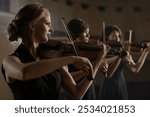 Team of three music players with classic wooden violins. Trio as band playing sounds of acoustic melody by bow close-up. Work of pretty women as orchestral violinists indoors at room of great theater