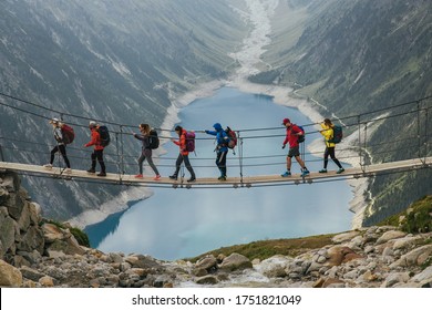 Team Of Travelers With A Backpack In The Mountains. A Group Of Travelers Crosses A Suspension Bridge Against The Backdrop Of A Mountain And A Glacier. Travel And Active Life Concept.
