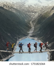 Team Of Travelers With A Backpack In The Mountains. A Group Of Travelers Crosses A Suspension Bridge Against The Backdrop Of A Mountain And A Glacier. Travel And Active Life Concept.