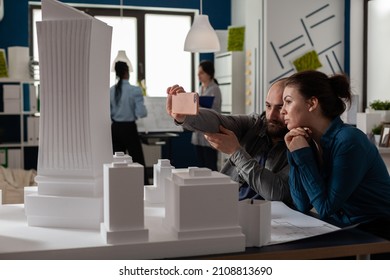 Team Of Two Architect Colleagues Using Smartphone In Videocall Conference With Client Presenting Desk With Buildings Models. Architectural Engineers Working Together With Smart Phone And Maquette.