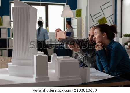 Team of two architect colleagues using smartphone in videocall conference with client presenting desk with buildings models. Architectural engineers working together with smart phone and maquette.