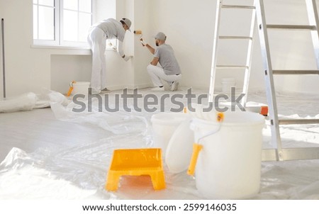 Team of two men construction workers painters painting walls with paint rollers, doing house repair or renovation, posing in empty room with equipment, working on interior design going to finish task 