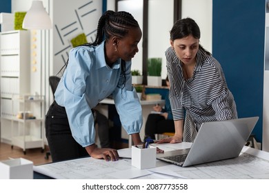 Team Of Two Professional Women Architects Working On Laptop Computer With Construction Plans On Desk In Architectural Office. Engineers Designing Blueprints For Real Estate Project.