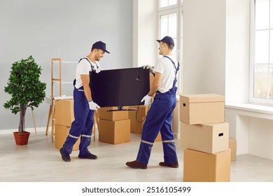 Team of two young cheerful professional workers of moving service in overalls carrying and delivering TV in new apartment with cardboard boxes. Move, moving day and relocation concept. - Powered by Shutterstock