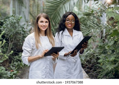 Team of two young pretty multiracial women botanists scientists, working in greenhouse with tropical plants and palm trees, smiling to camera with clipboard and tablet - Powered by Shutterstock