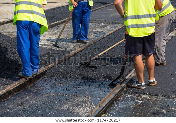 Team of\
workers put the hot asphalt on a street along tram car\'s railroad\
lines. Road construction workers with shovels in protective\
uniforms. Working in the hot\
day.