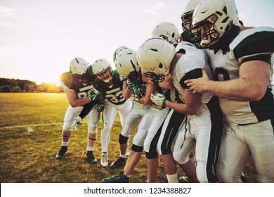 Team Of Young American Football Players Having Fun Trying To Grab A Football From Their Captain While Standing On A Sports Field During A Practice