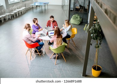 Team Of A Young Coworkers Dressed Casually Working Together With Laptops Sitting At The Round Table In The Office, Wide View On The Office From Above