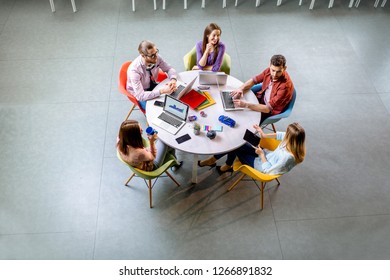 Team Of A Young Coworkers Dressed Casually Working Together With Laptops Sitting At The Round Table In The Office, View From Above