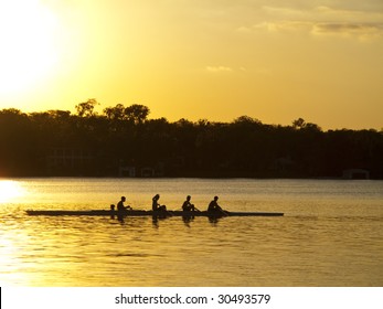 A Team Of Young Mean In A Row Boat Silhouetted Against Setting Sun