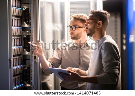 Team of young multi-ethnic specialists in glasses standing at server and using tablet while managing network server together in data center