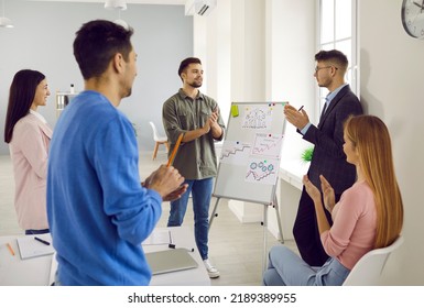 Team Of Young People Having A Work Meeting And Using An Office Whiteboard. Group Of Employees Applauding After Their Coworker Has Made A Good Business Presentation With Graphs And Other Visuals