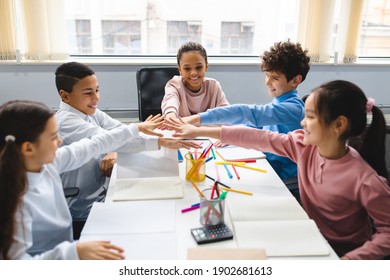 Teamwok And Children Concept. Diverse excited group of emotional happy junior school kids sitting at desk in classroom and putting hands together, having fun, studying or playing game