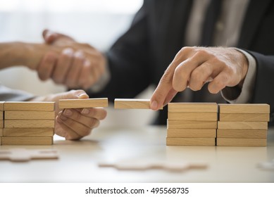 Teamwork or building bridges concept with a businessman and woman holding wooden building blocks to form a bridge over a gap while clasping hands in the background.