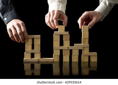 Teamwork And Collaboration - Hands Of Three Businessman Assembling A Balanced And Stable Structure Of Wooden Cubes On A Black Desk With Reflection And Black Background.