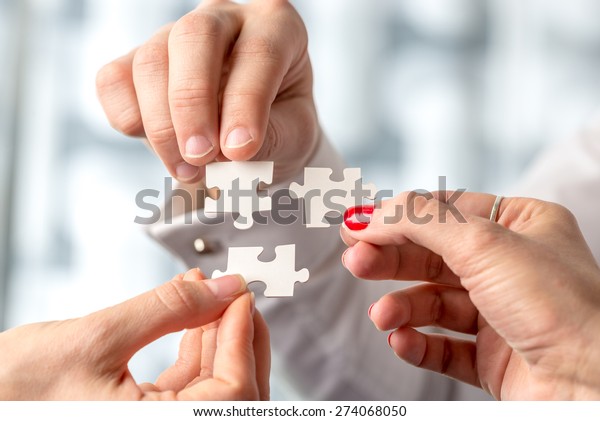 Teamwork concept using white puzzle pieces being\
fitted together by three male and female hands in a challenge,\
brainstorming and solution\
concept.