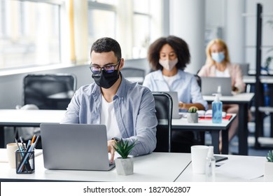 Teamwork In Corporate Company And Returning To Work After Quarantine Covid-19. Focused Millennial Man In Glasses And Protective Mask Works At Laptop At Workplace With Antiseptic In Office Interior