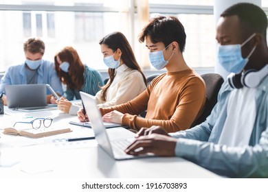 Teamwork in corporate company and returning to work after quarantine covid-19. Focused millennial people wearing medical masks working using laptop and tablet, writing at workplace in office interior