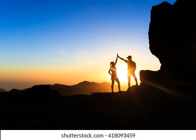 Teamwork Couple Helping Hand Trust Help, Silhouette Success In Mountains. Team Of Climbers Man And Woman. Hikers Celebrate With Hands Up, Help Each Other On Top Of Mountain, Happy Climbing Together.