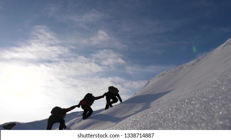 Teamwork Desire To Win. Climbers Derajat Each Other's Hands To Help A Friend Climb To Top Of Snowy Mountain. Travelers In Winter On A Hill In Bright Rays Of The Sun. Concept Of Sports Tourism.