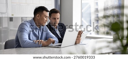 Teamwork on project development. Wide panoramic web banner image two diverse male business coworkers sit together at office table compare data on laptop tablet synchronize information coordinate tasks