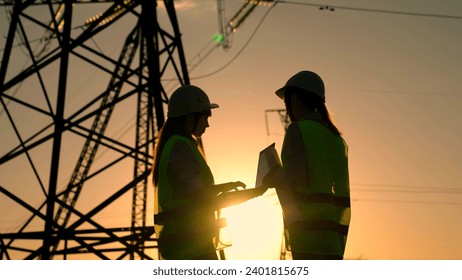 Teamwork of power engineers with computer in protective helmets, maintenance of power lines in outdoors. Two Female civil engineers work together using computer on power line. Green energy. Builders
