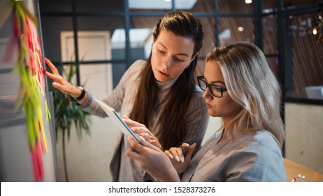 Teamwork, project management, agile methodology. Two young business women in the office are working on a startup. Colleagues plan work and stages of development of company, glue sticky leaves on Board