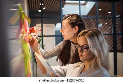 Teamwork, project management, agile methodology. Two young business women in the office are planning product development and support. Colleagues glue sticky stickers on the Board - Shutterstock ID 1528376318