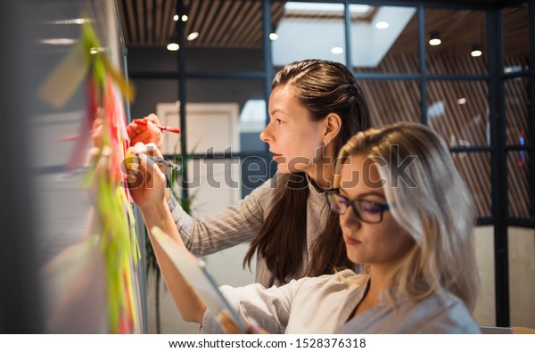 Teamwork, project\
management, agile methodology. Two young business women in the\
office are planning product development and support. Colleagues\
glue sticky stickers on the\
Board