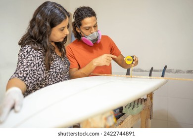 Teamwork with protective work gear using tape to measure a surfboard in a workshop - Powered by Shutterstock