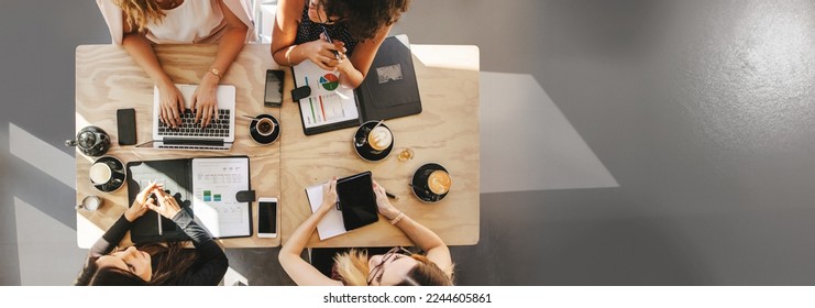 Teamwork in a small business team of women entrepreneurs discusses startup ideas. Top view of four women sitting around table in cafe with laptop, digital tablet and documents. Group of women working  - Powered by Shutterstock