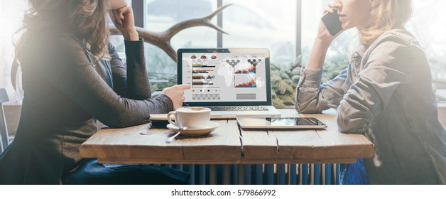 Teamwork, two young businesswomen sitting at table. On table laptop, coffee cup and tablet computer. First girl showing pencil on graphs and charts on computer screen, other is talking on cell phone. - Powered by Shutterstock