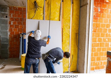 Teamwork, workers are measuring with a tape measure, checking wall dimensions before installing the plasterboards on the partition insulated wall. Building is under construction.