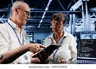 Teamworking colleagues updating server racks, writing intricate binary code scripts on tablet and laptop. Elderly coworkers using programming to fortify cybersecurity in high tech workspace