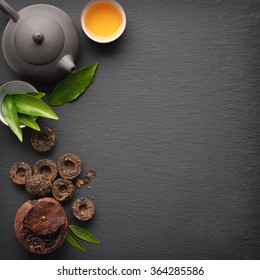Teapot with cup and dry green tea pu-erh on black slate background, top view