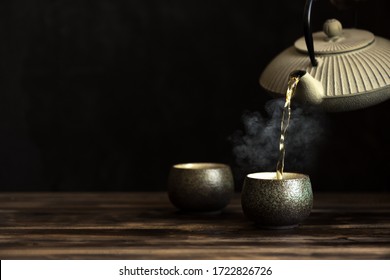 Teapot Pouring Tea Into Cups. Traditional Japanese Chinese Iron Teapot And Ceramic Teacups, Asian Tea Set On Dark Background, Copy Space.