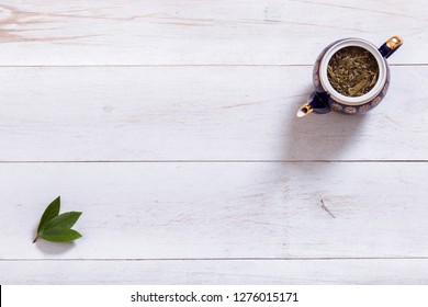 Teapot And Tea Time Leaves Set On White Wooden Table, Black Herbal Homemade Hot Beverage In Porcelain Pot With Green Leaf, English Tea Ceremony Background, Top View From Above, Copy Space